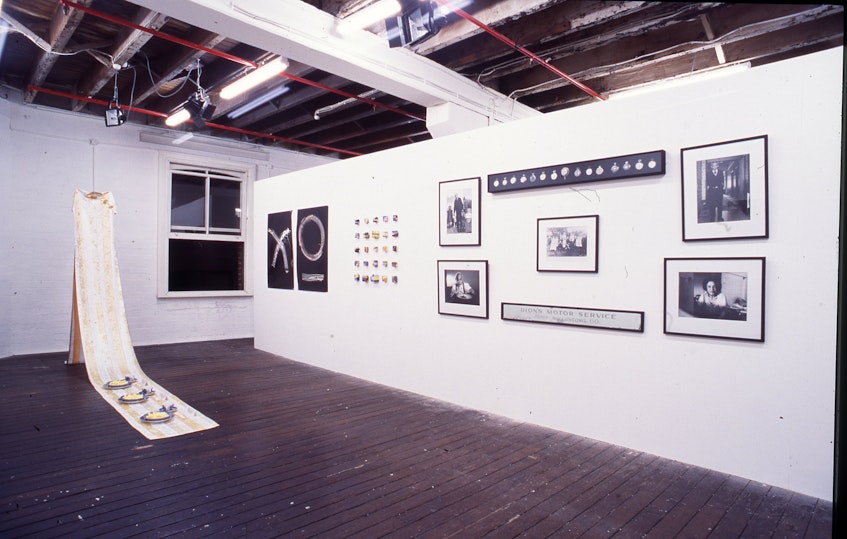 L-R: Tiffany Lee-Shoy, Untitled, 1999, hand stitched cheong-sam, Chinese embroidered satin, lurex thread, huaniu satin binding, willow pattern china, digitally printed tracing paper and banana lollies; Montri Muenouy, In Remembrance of, 1993, B+W photograph; Ngoc Tran, Untitled, 1999, wax and oil paint on velvet jewellery box; Tom Dion, Barney, 1993, silver gelatin print; Tom Dion, Ernie, 1993, silver gelatin print; Tom Dion, Watches, 1999, wood, glass, velvet, pins, watches, On loan from the Charles Dion Archive; Tom Dion, The Dion Family, photo taken 1919, resin coated print, On loan from the Charles Dion Archive; Tom Dion, Bus Mirror, circa 1940, mirrored glass and wood, On loan from the Charles Dion Archive; Tom Dion, Uncle Tom, 1994, silver gelatin print, On loan from the Charles Dion Archive; Tom Dion, Alma, 1993, silver gelatin print, On loan from the Charles Dion Archive.