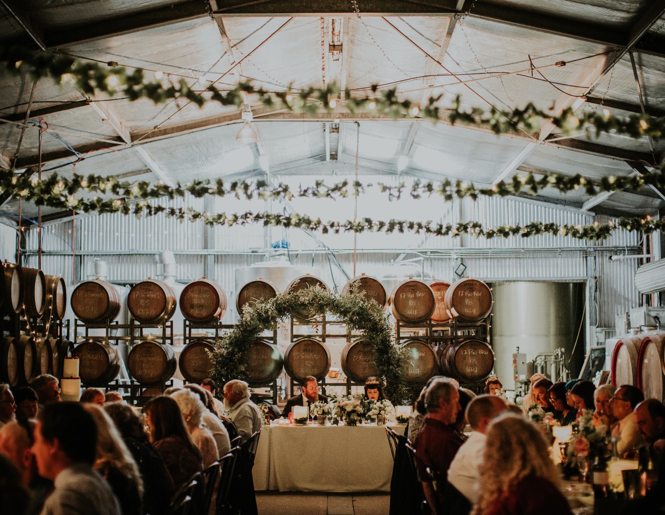 Couple sitting at wedding party table, with wine barrel racks behind them.