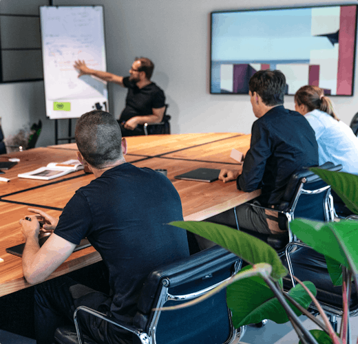 People sitting around meetingroom table watching presenter explain on whiteboard
