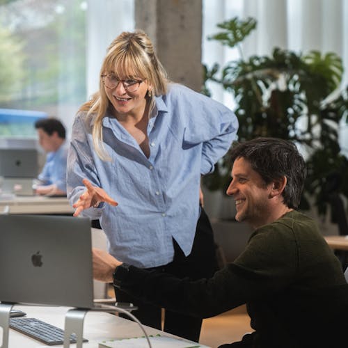 A female Craftzing colleague with a blue shirt and long blond hair is discussing something with a male Craftzing colleague with dark brown hair and a dark green sweater. Together they laugh while looking at a laptop screen.