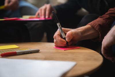 A close-up of a hand writing on a bright pink post-it with a black marker