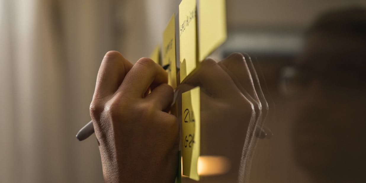 A hand writing on a post-it with a black marker while it hangs on a window with other post-its