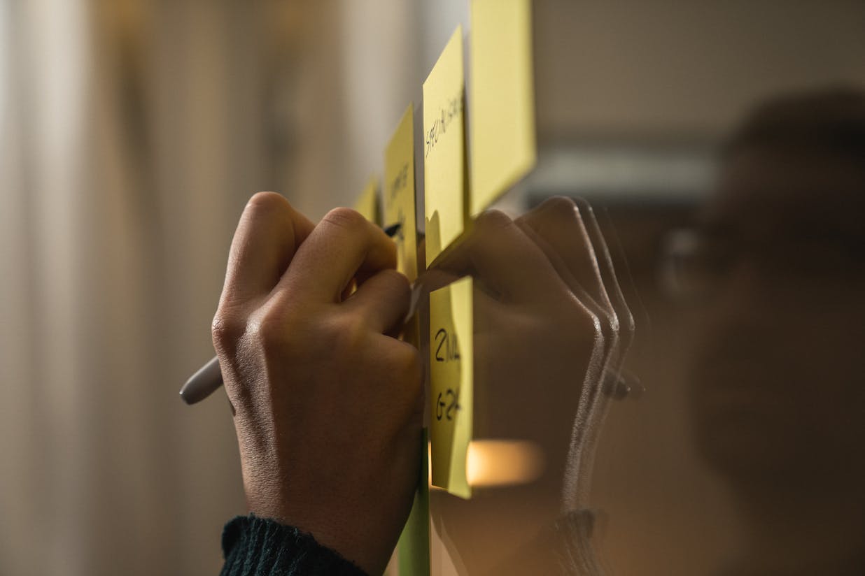 A hand writing on a post-it with a black marker while it hangs on a window with other post-its
