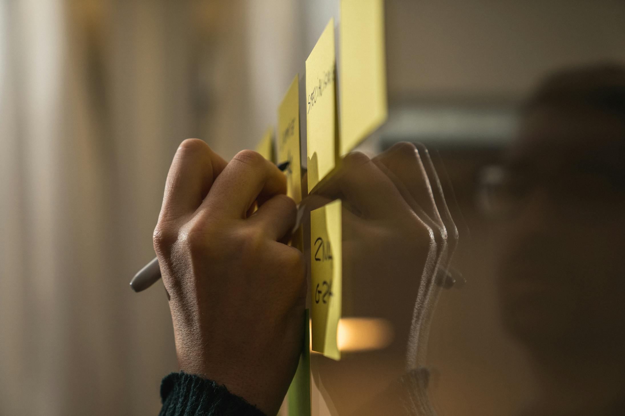 Een hand die met een zwarte stift op een post-it schrijft terwijl deze op een raam hangt met andere post-its