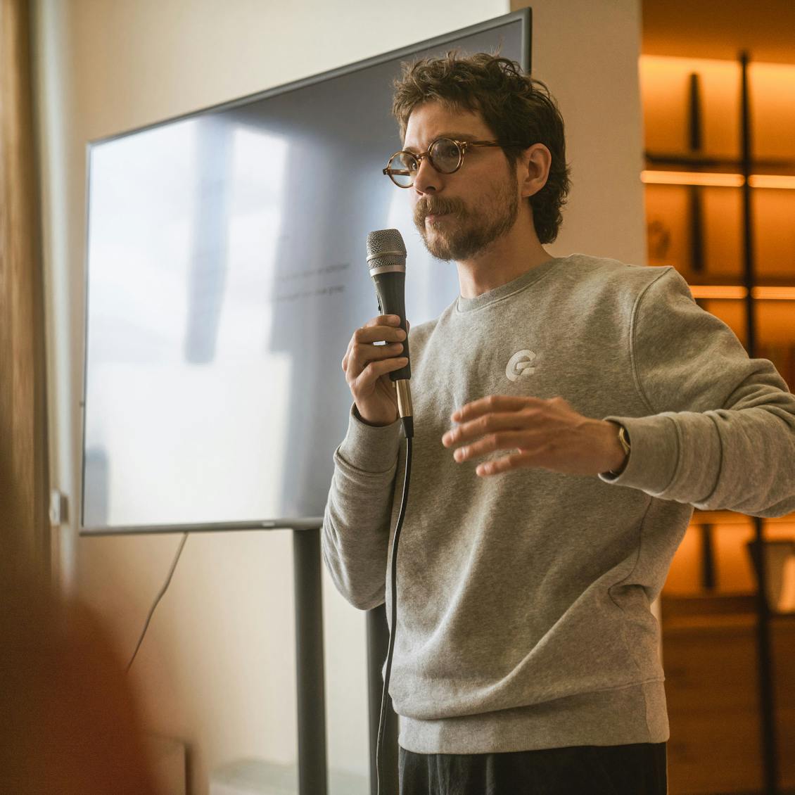 Man in a grey sweater holding a microphone explaining something with a big tv screen in the back.