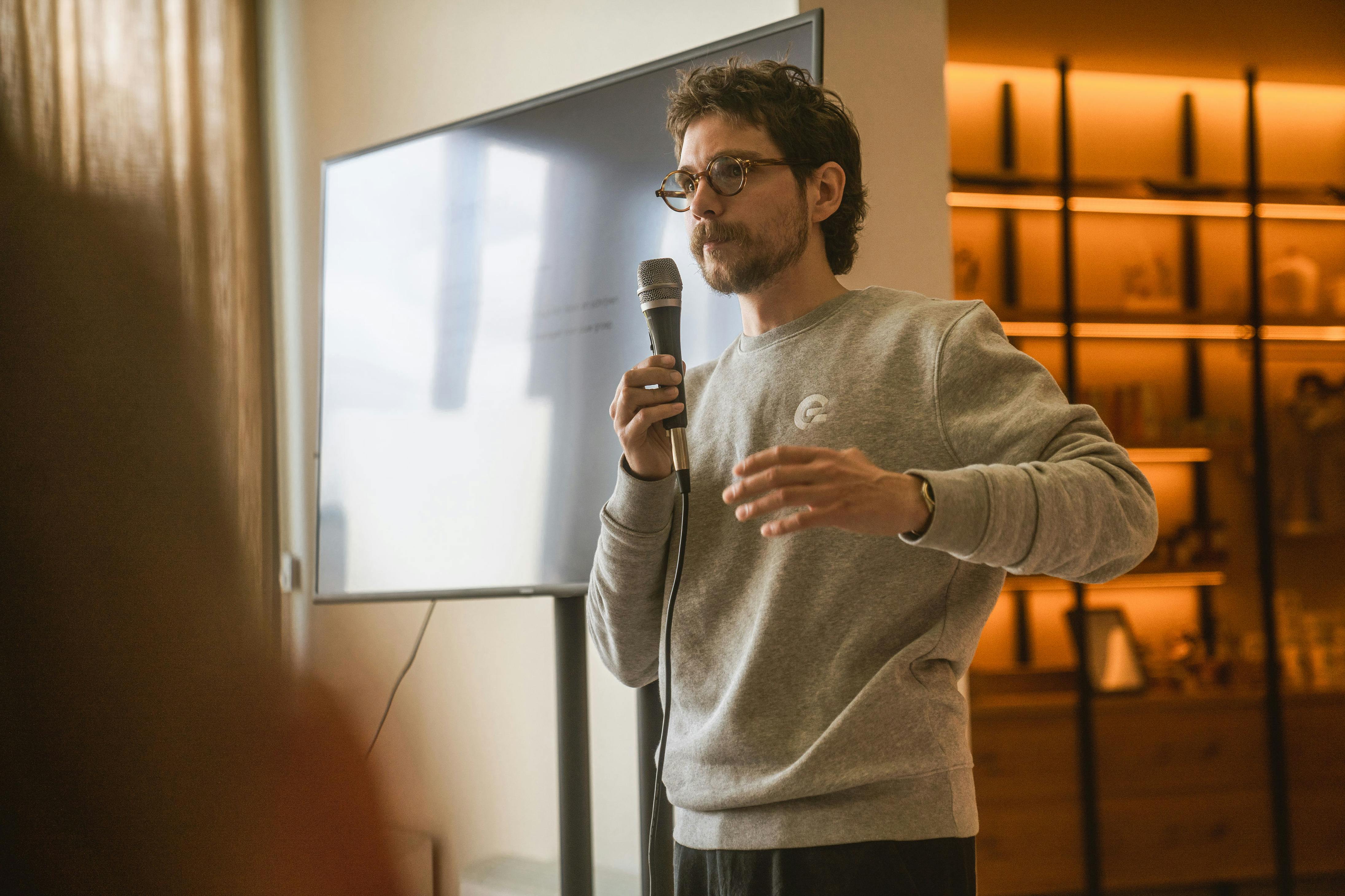Man in a grey sweater holding a microphone explaining something with a big tv screen in the back.