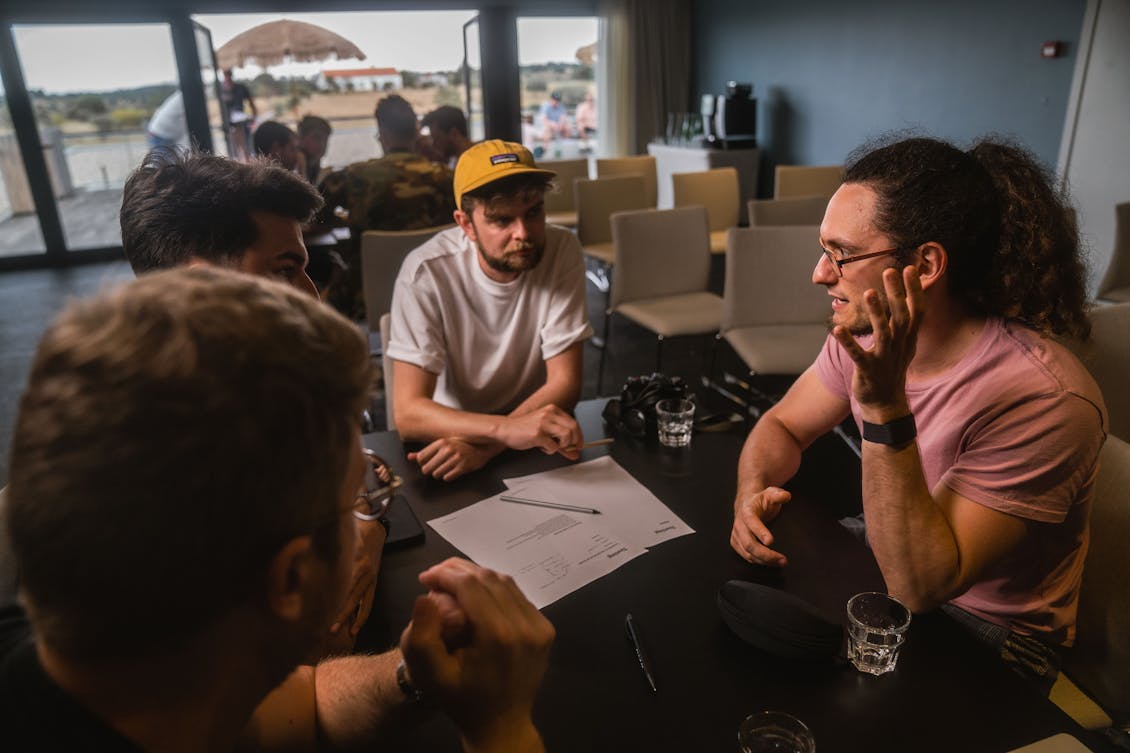 Four men sitting at a table discussing something. In the background another group of people sitting at a table and discussing something.