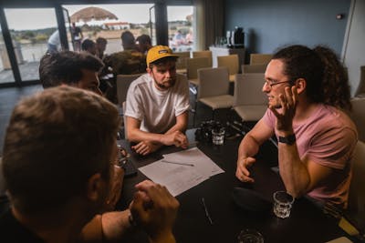 Four men sitting at a table discussing something. In the background another group of people sitting at a table and discussing something.