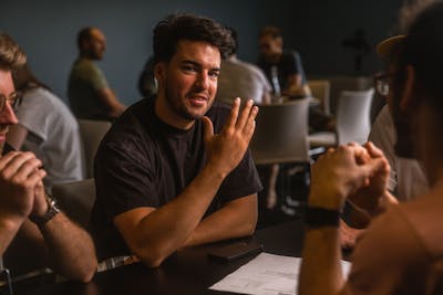 Close-up of a man with dark hair explaining something to a group of other people at a table while raising his hand.