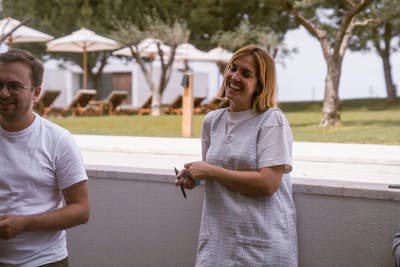 Woman wearing a white dress laughing while leaning on to a wall. Left of her a man with a white t-shirt and glasses with a smile on his face.  In the background you see the garden of a hotel with sunbeds and umbrellas.