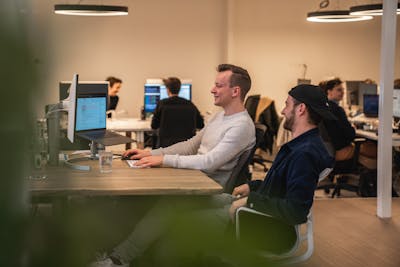 2 men sitting behind a desk, one of them behind his computer, the other one next to him, both looking a the screen. Some other people working behind their computers in the background.