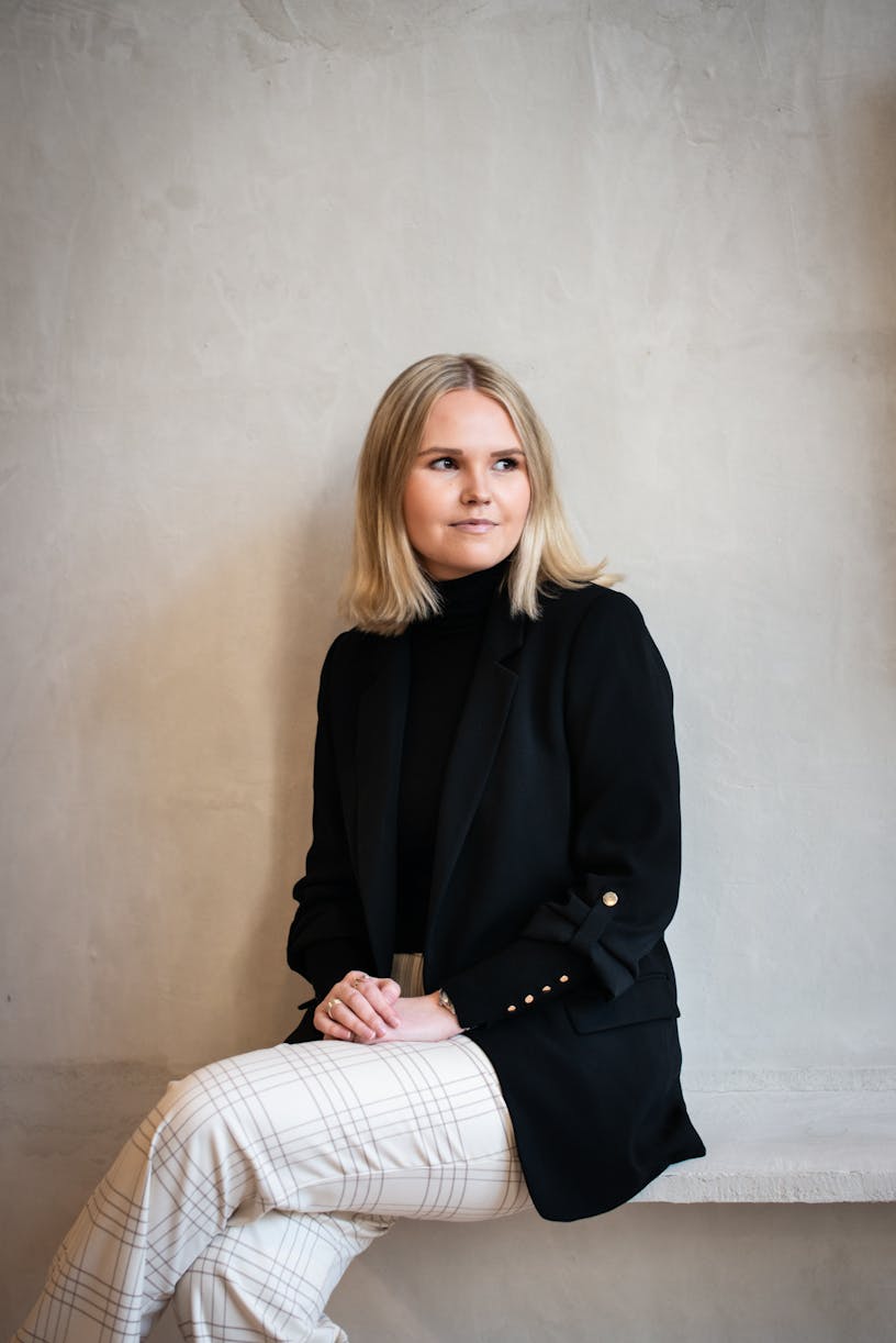 Business analyst Fien Boone sitting on a bench. Fien is wearing a black turtle neck sweater, a black blazer and has blond shoulder length hair.