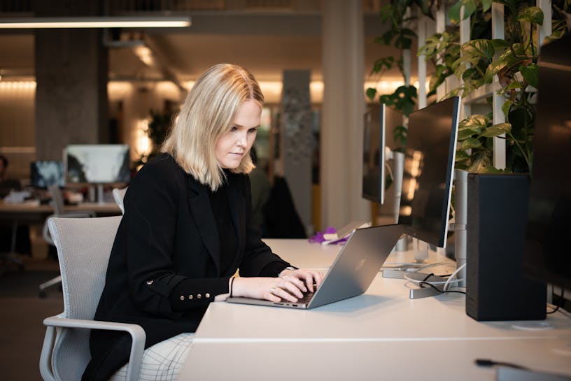 Business analyst Fien Boone sitting at a desk behind her laptop. Fien is wearing a black turtle neck sweater, a black blazer and has blond shoulder length hair.