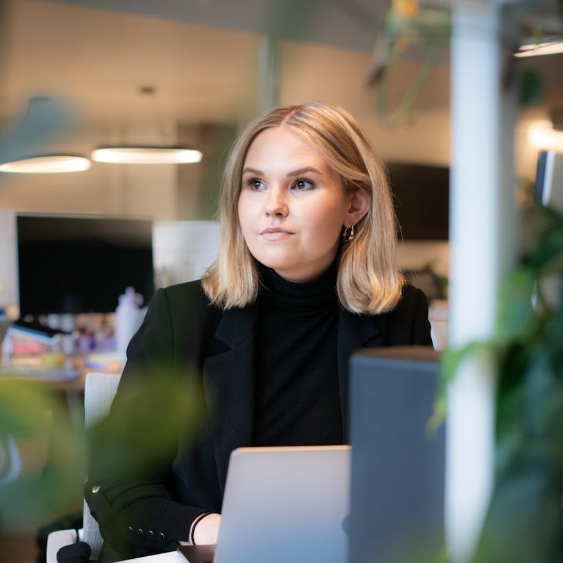 Business analyst Fien Boone sitting at a desk behind her laptop. Fien is wearing a black turtle neck sweater, a black blazer and has blond shoulder length hair.