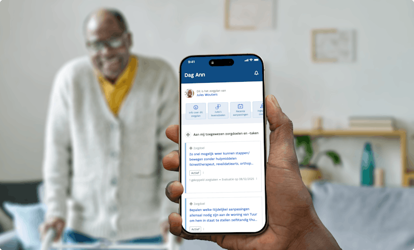 Hand holding an Iphone that shows the interface of Alivia . In the background an older man of colour with glasses, a yellow shirt and a beige vest is standing in his living room, leaning against a rollator and smiling in the camera.