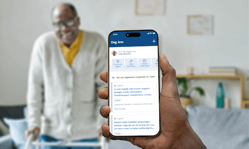 Hand holding an Iphone that shows the interface of Alivia . In the background an older man of colour with glasses, a yellow shirt and a beige vest is standing in his living room, leaning against a rollator and smiling in the cmaera.