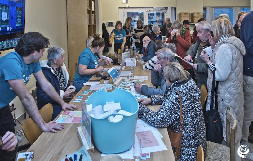 Group of people standing around a large table at meeting center KomAf. Some older people seek support from 'Digihelpers'.