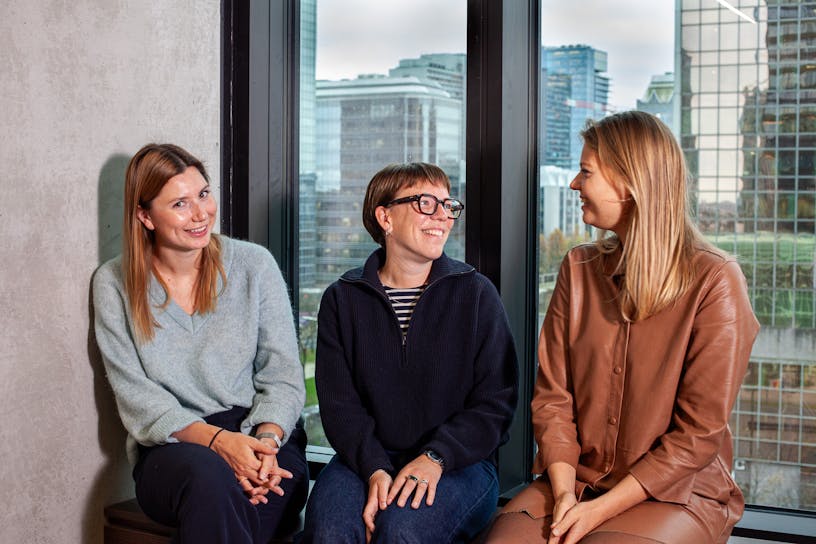 Group picture of Camille Vanremoortele, Laura Peeters and Hanne Verheust. They're sitting next to each other and smile at each other.
