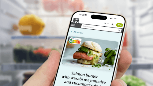 Iphone mock-up of a recipe page for a salmon burger on the Ahold Delhaize app. You see a hand holding the Iphone and a filled fridge in the background.for a salmon burger
