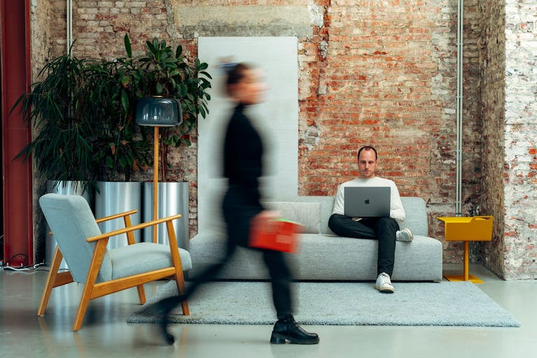 Craftzing colleague sitting in a grey sofa in Craftzing Ghent office with his laptop on his laptop while another colleague passes by.