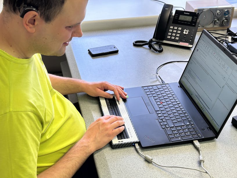 Stijn Van Roey, a person with a visual impairment, sits at a desk and behind is laptop and is using digital applications with the help of his screenreader.