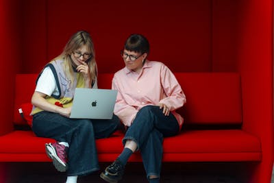 Two female Craftzing colleagues sit together on a red bench, watching something on one of their laptop screens.