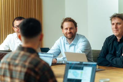 Three male Craftzing colleagues sit at a wooden desk in a meeting room, attentively listening to someone across from them whose face is not visible.
