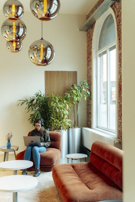 A woman sits in an orange couch in the office lobby, wearing headphones and working on her laptop. Next to her is a larger version of the same couch, and behind her are green plants and large round modern lamps lighting the space.