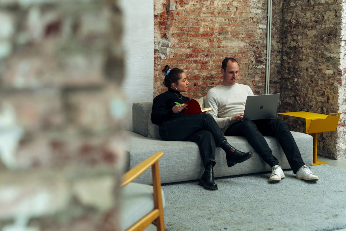 A male and female colleague sit on a grey sofa. He shows something on his laptop while she, holding a pen and orange booklet, watches and listens.