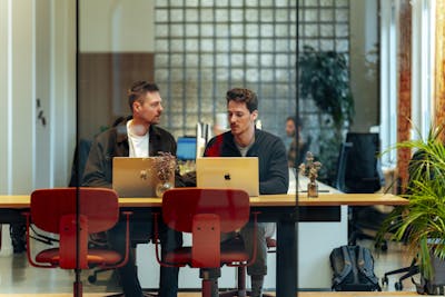 Two male Craftzing colleagues sit at a long wooden table, discussing something with their laptops in front of them. Two empty chairs are across from them. The background shows an urban office with brick walls, glass, and green plants.