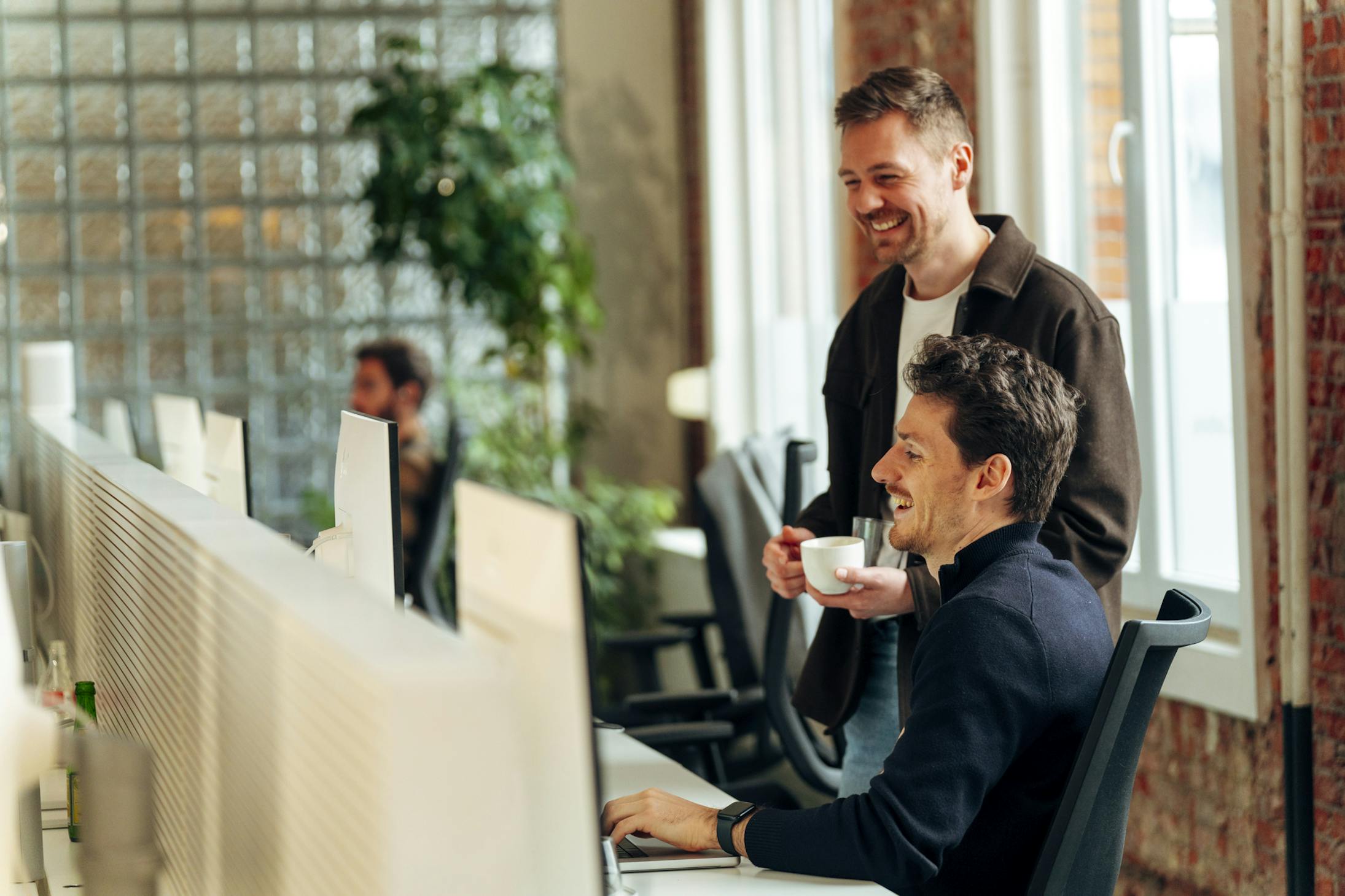 Two casually dressed men, one sitting behind a desk and the other standing beside him, laughing while looking at a laptop screen, seemingly reacting to something humorous.
