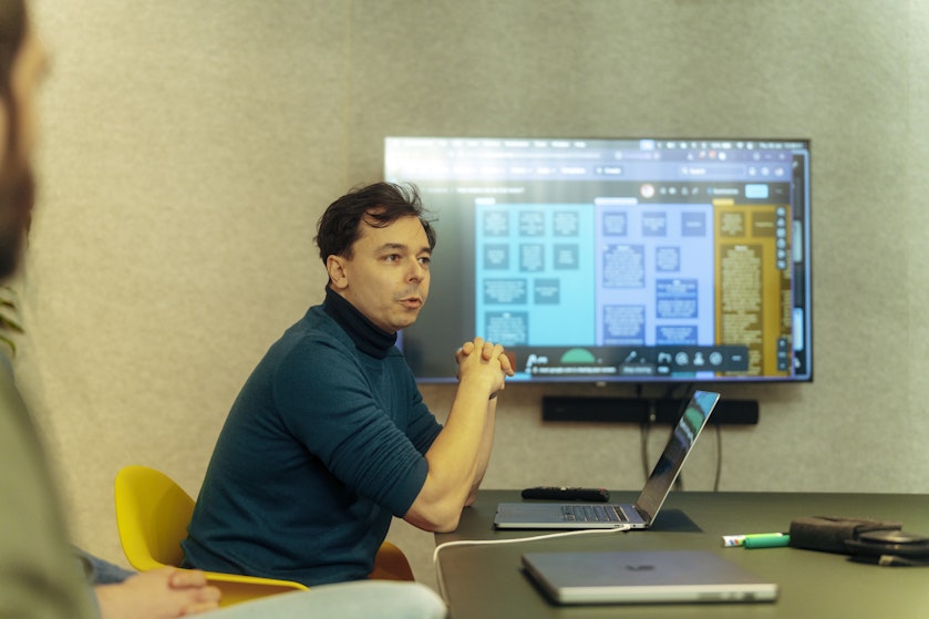 Man with dark hair and a dark blue shirt leads a presentation from his laptop, using a large screen behind him to address a group of people.