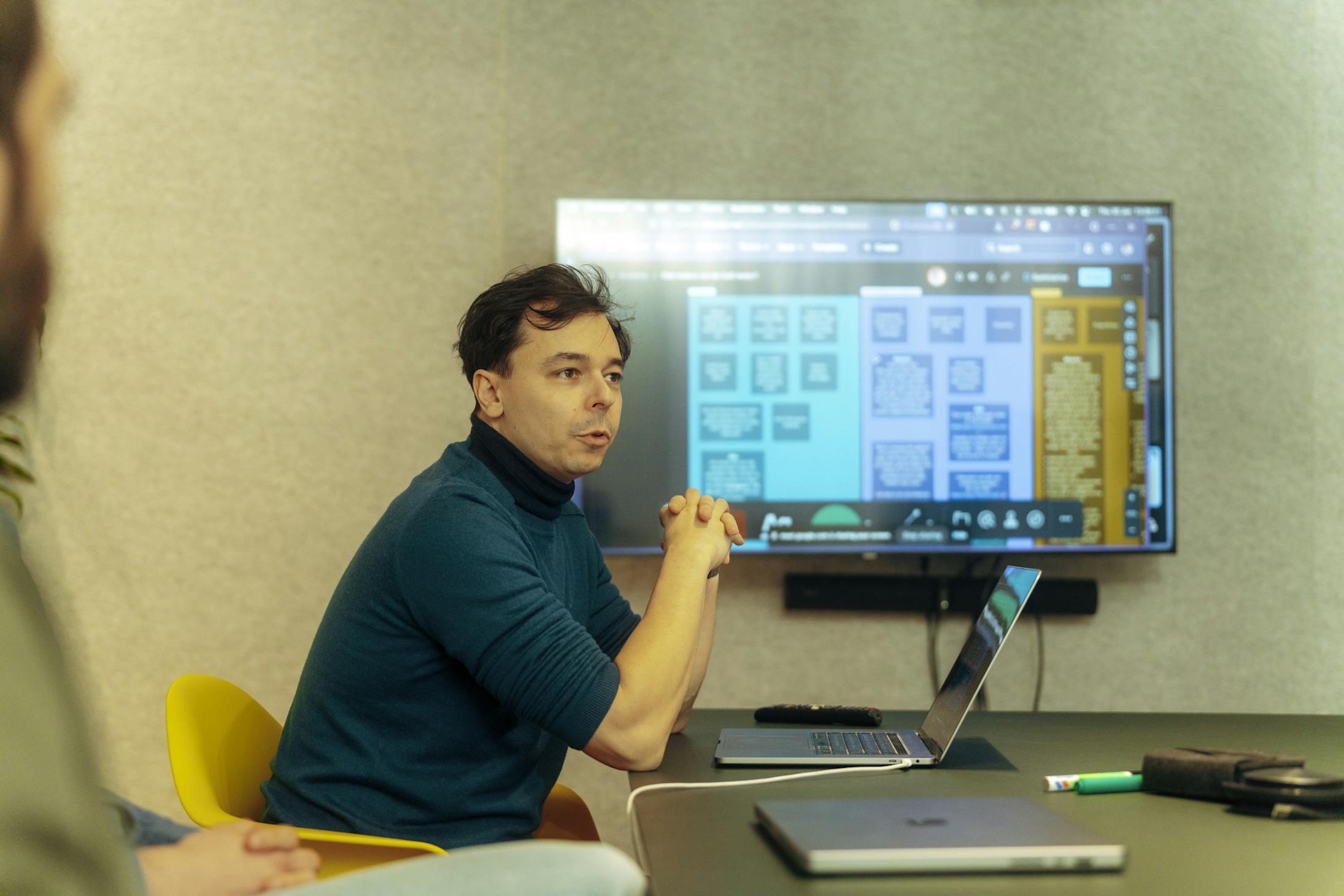 Man with dark hair and a dark blue shirt leads a presentation from his laptop, using a large screen behind him to address a group of people.