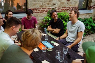 Seven Craftzing colleagues—six men and one woman—are gathered around a dark wooden table outside, playing a game. One of them bursts out laughing as the others look on with amusement.