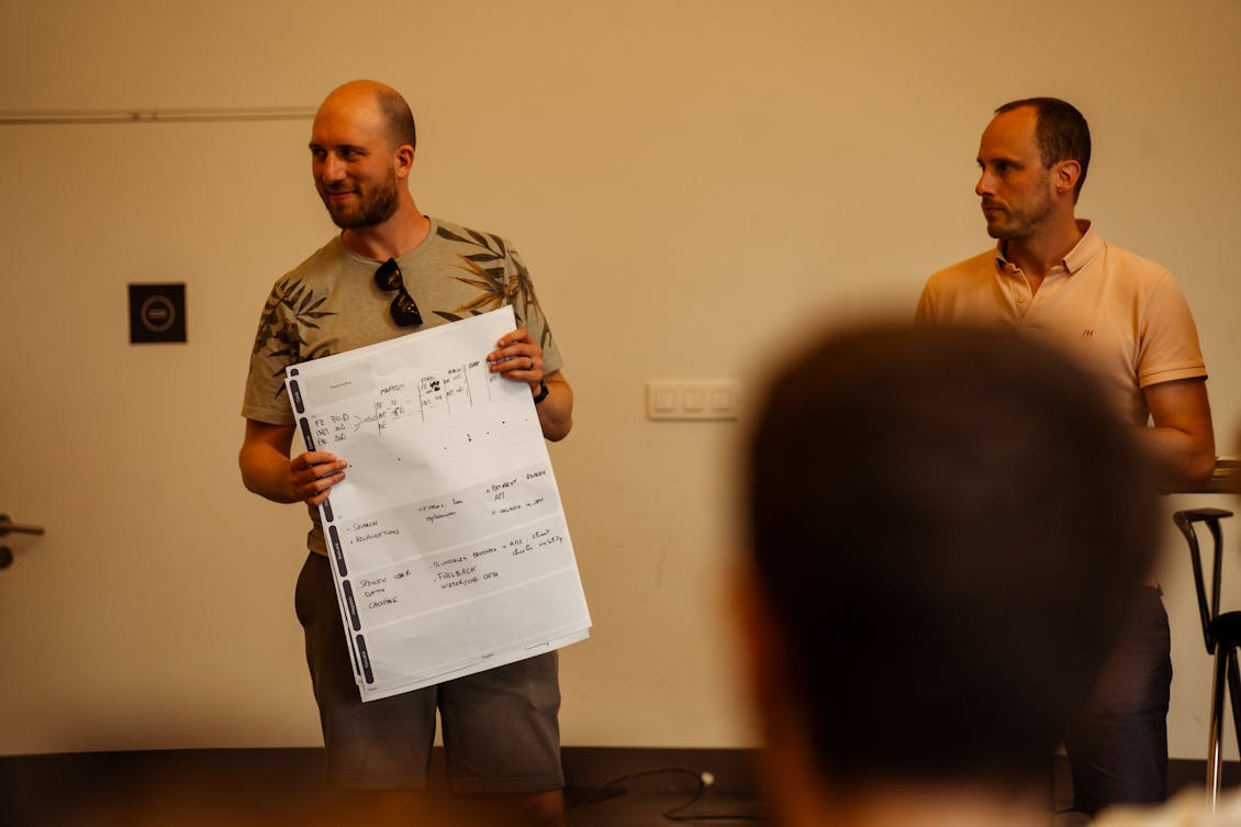 Two men stand side by side, one holding a large paper and presenting to an unseen audience in front of them.