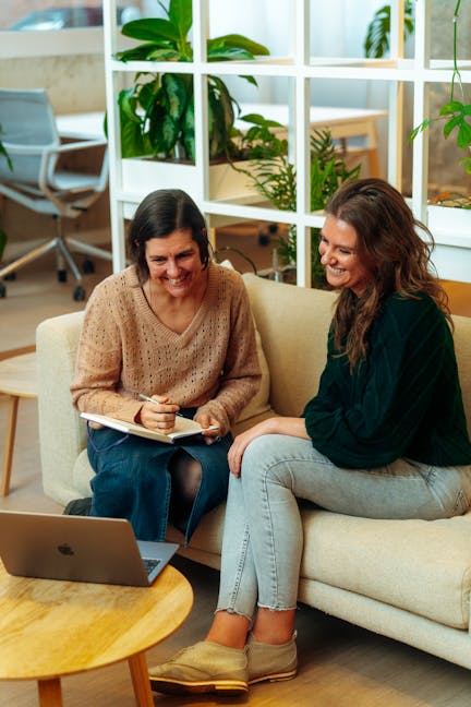 Seated side by side on a beige sofa, two female colleagues focus on a laptop in front of them as they discuss something. A lush plant wall forms the backdrop.