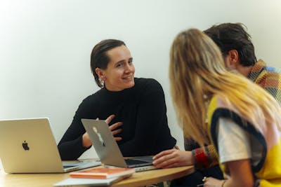 A woman sits at a round table behind her laptop, explaining something to a man and a woman whose faces are not visible.