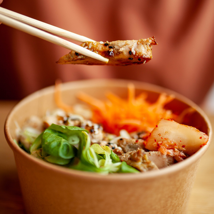 teriyaki chicken donburi in a takeaway container with a piece of chicken held above it with chopsticks