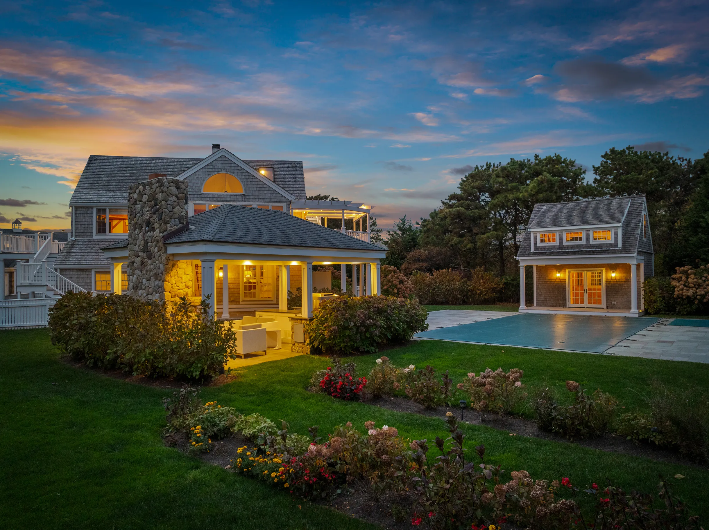 A property in Massachusetts at sunset. Two grey houses, both with lights on, and a garden and pool in front of them.