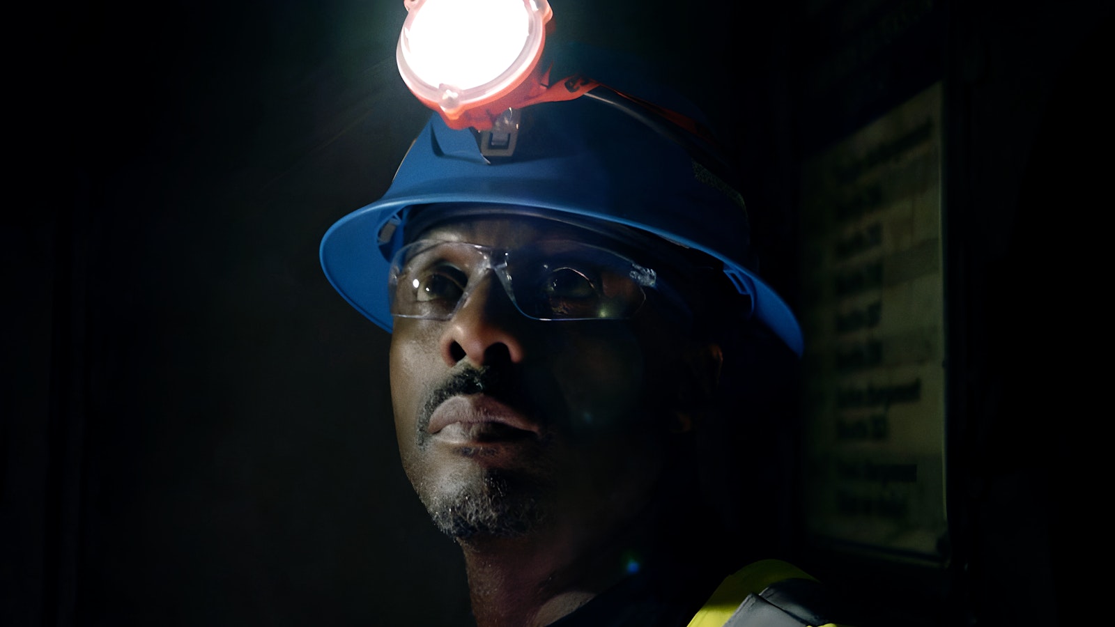 Idris Elba looks into the dark at LaRonde Complex wearing a blue hard hat fitted with a safety lamp.
