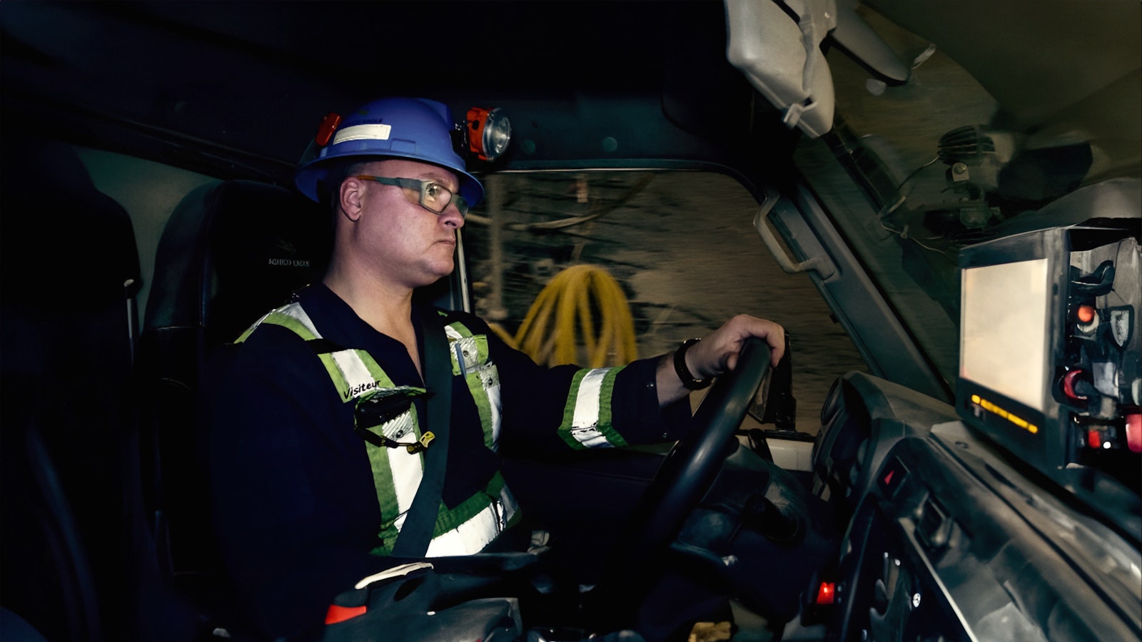 A miner sits driving a truck underground. In the cab he wears a blue safety hat with a light and fluorescent yellow stripes.