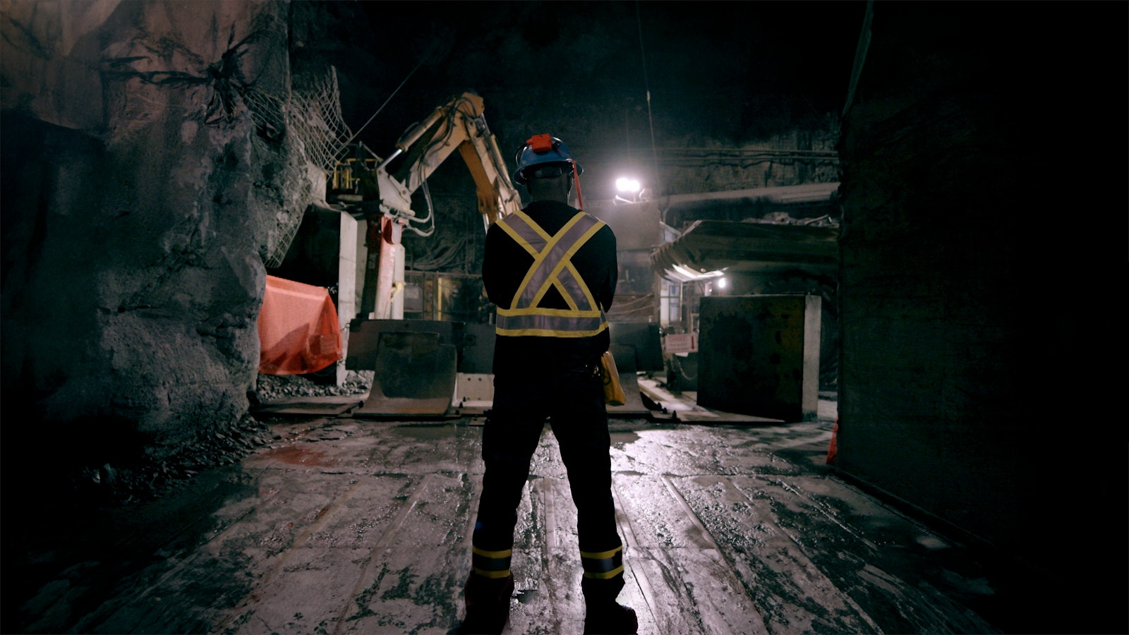 A miner stands in dark blue safety gear with yellow visibility sashes in front of the mine’s machinery.