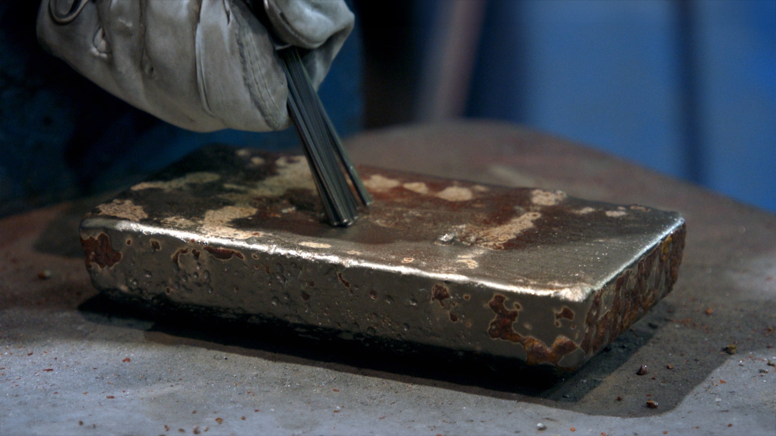 A miner’s glove holds a large grey metal brush and cleans some of the smelting residue from a newly cooled gold bar.