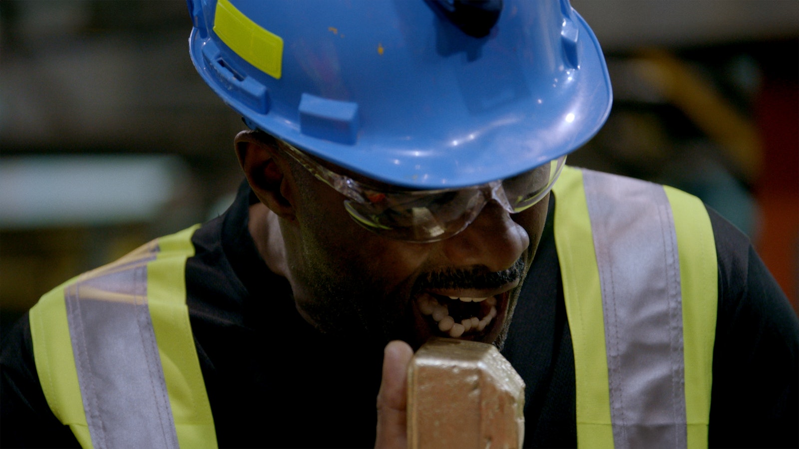 Wearing a blue safety hat and yellow jacket, Idris Elba holds up a large gold bar and pretends to bite into it.