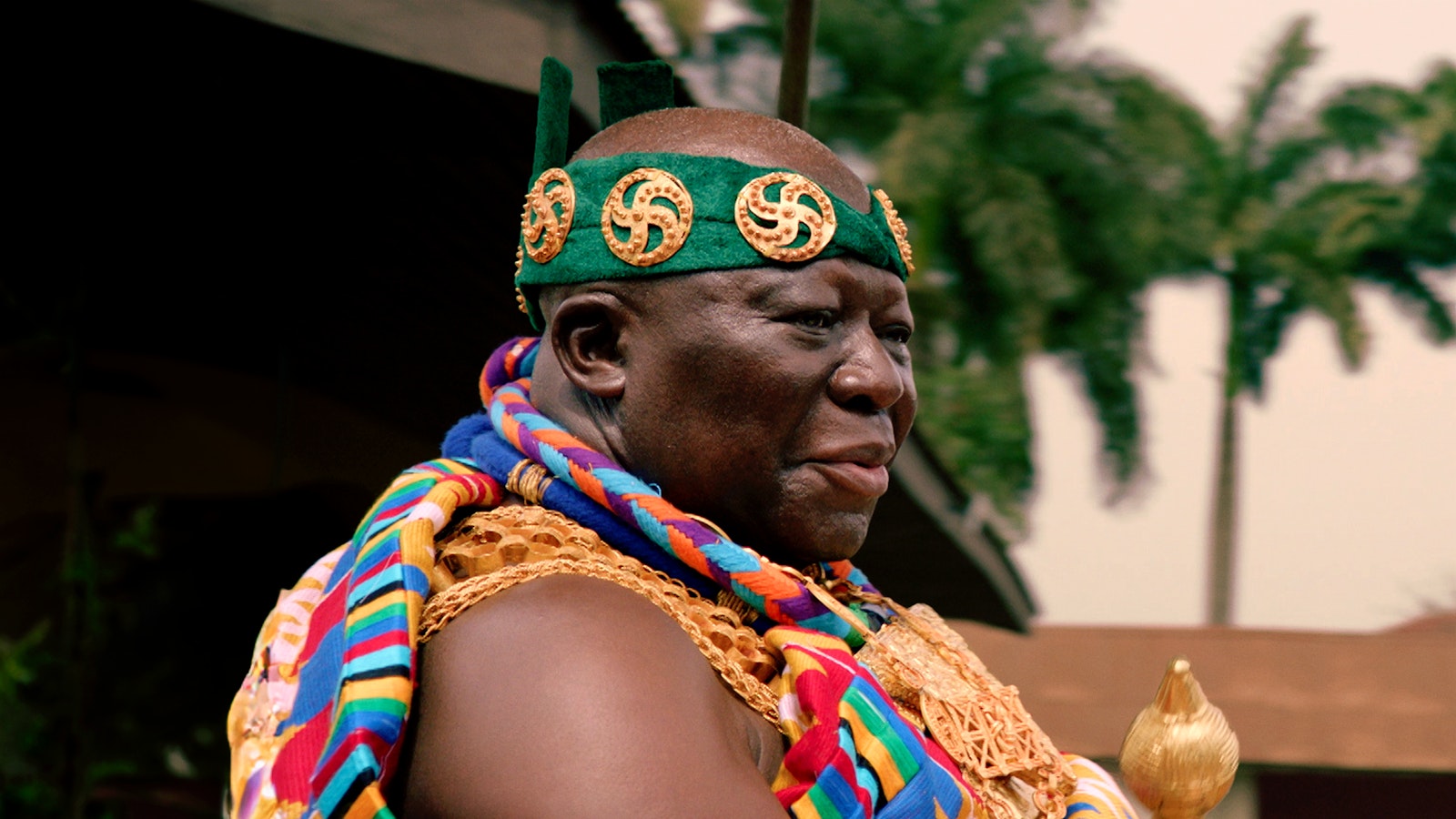 A photo portrait of Ghana's Ashanti King who wears many gold adornments among colourful robes and a green headdress.