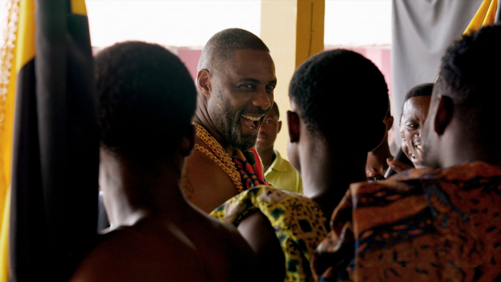 Idris Elba smiles and laughs while celebrating at the festival. In front of him we see the backs of two festival goers' heads.