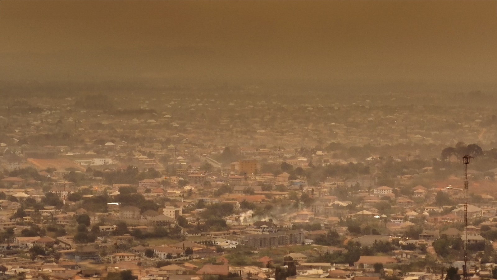 An aerial shot of the city of Kumasi, beneath the hot, orange, African sky extends as sprawling suburban metropolis.