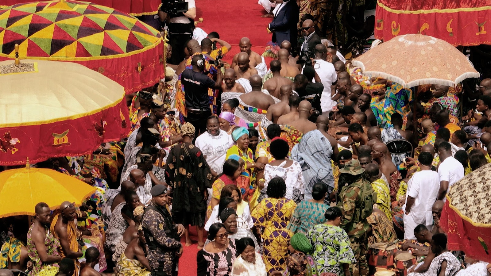 A closer shot of the red carpet leading to the king's throne. Large parasols and people in colourful robes line the way.