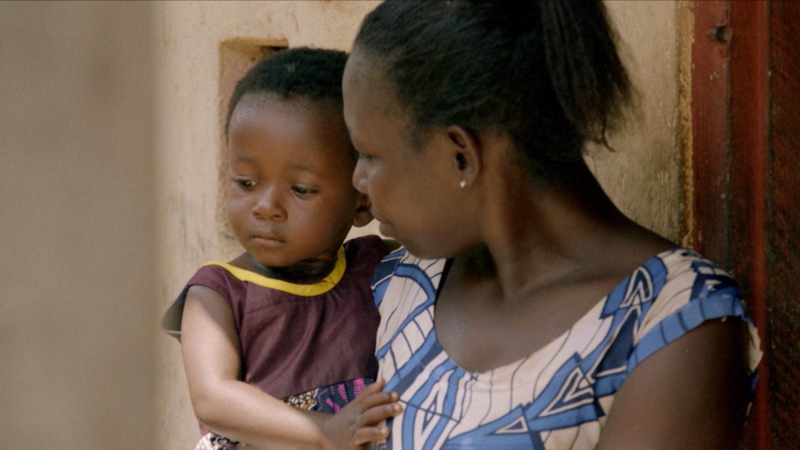 A mother holds her child in her arms. With an expression of approval, she looks at the child who is looking to their right.