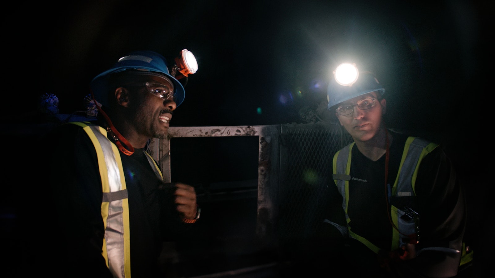 Idris Elba and miner Pascal Laroche look at one another wearing high visibility jackets and headlamps.